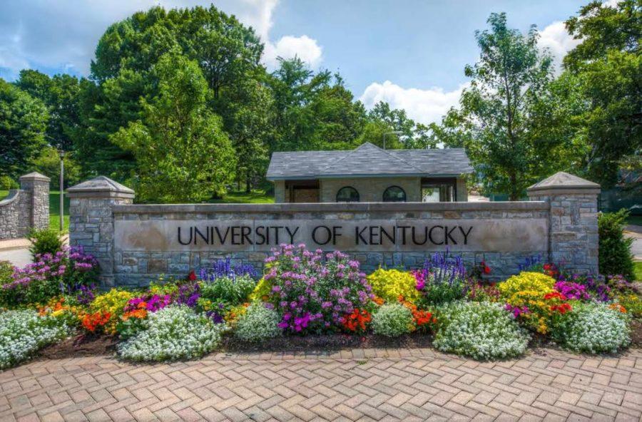 The entrance to the University of Kentucky's front campus near the Gatton School of Business and Economics and the Main Building is shown in spring. Landscaped flowering plants in purples, magenta, yellow, scarlett, orange and white are planted in front of the stone entrance marker that says "University of Kentucky." The sky is blue and the sun shines bright.